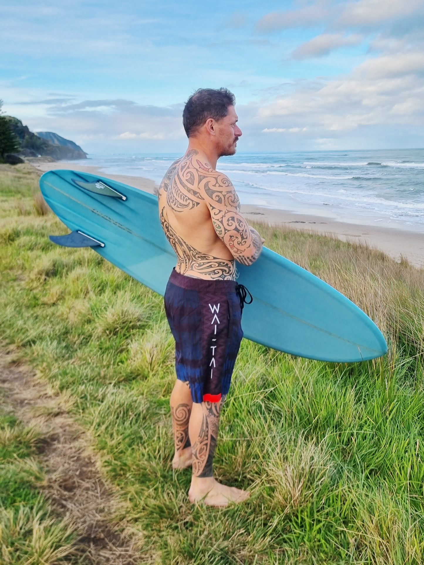 Wai-Tai Aotearoa Piu 4 Way Stretch Maori Boardshorts on Maia Gibbs carrying his Maori surfboard at Okitu Beach in Gisborne