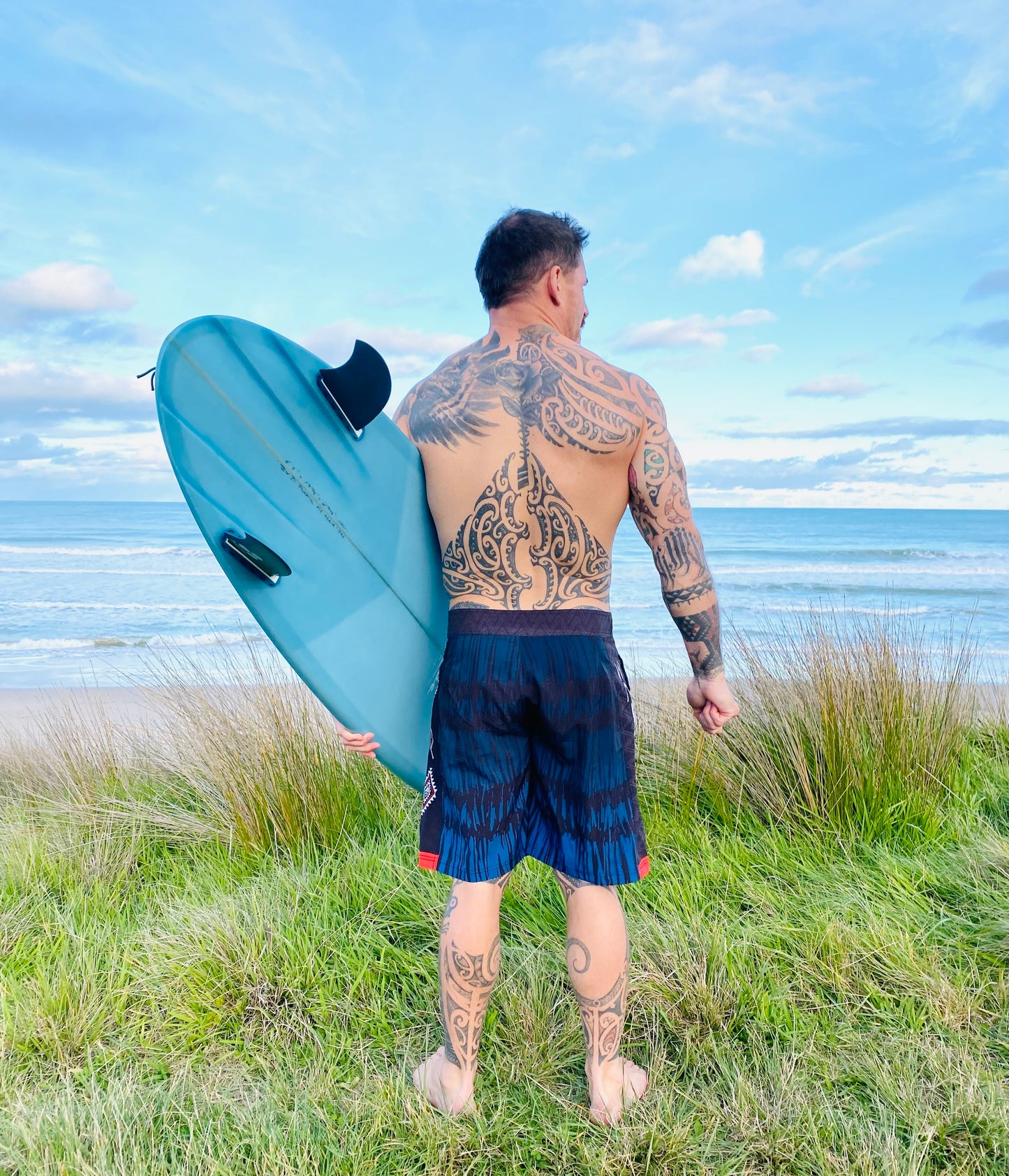 Wai-Tai Aotearoa Piu 4 Way Stretch Maori Boardshorts on Maia Gibbs holding his Maori surfboard at Okitu beach