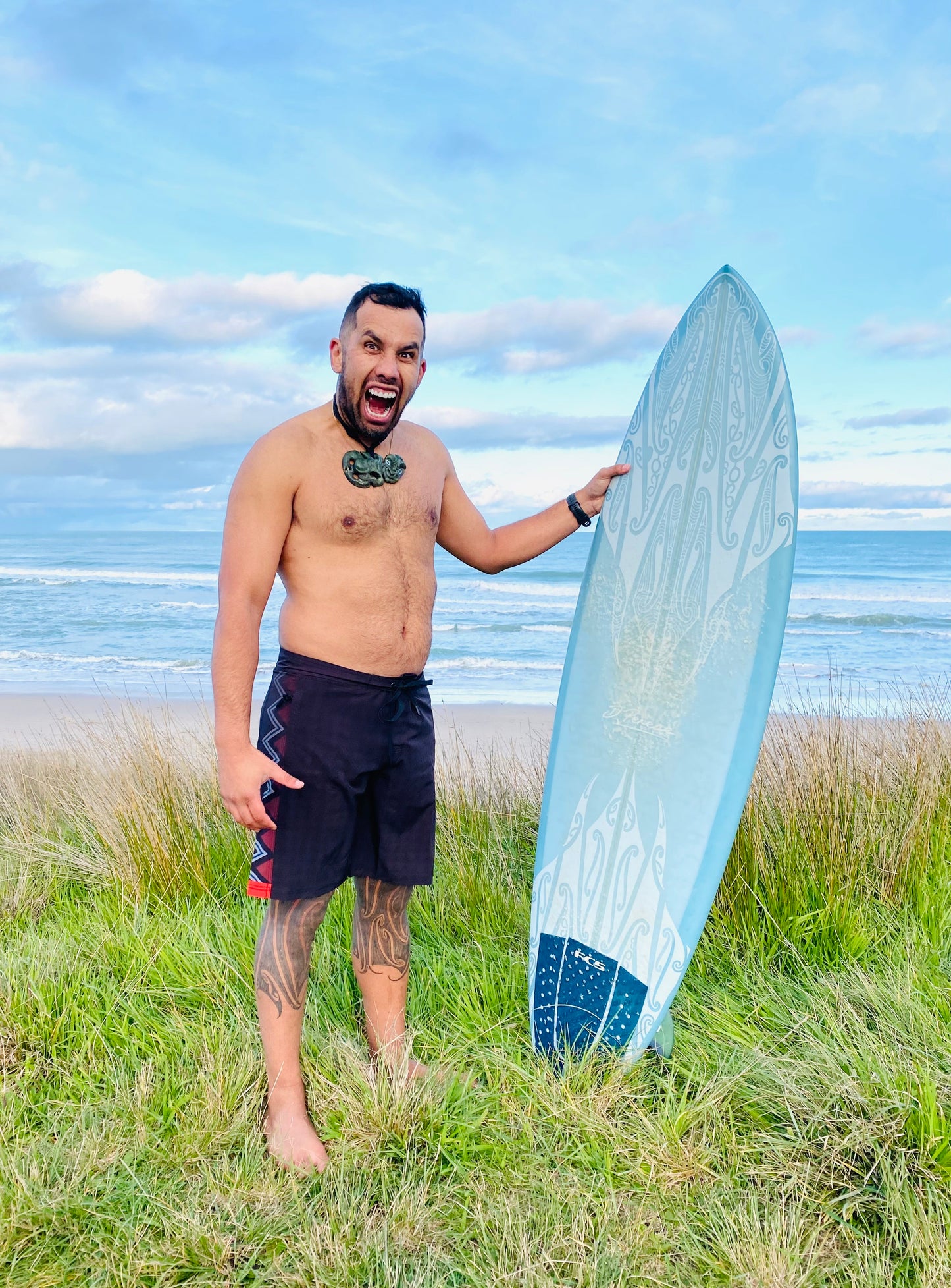 Wai-Tai Aotearoa Taniwha 4 Way Stretch Maori Boardshorts on Daniel Procter holding a Maori design surfboard from front at Okitu beach
