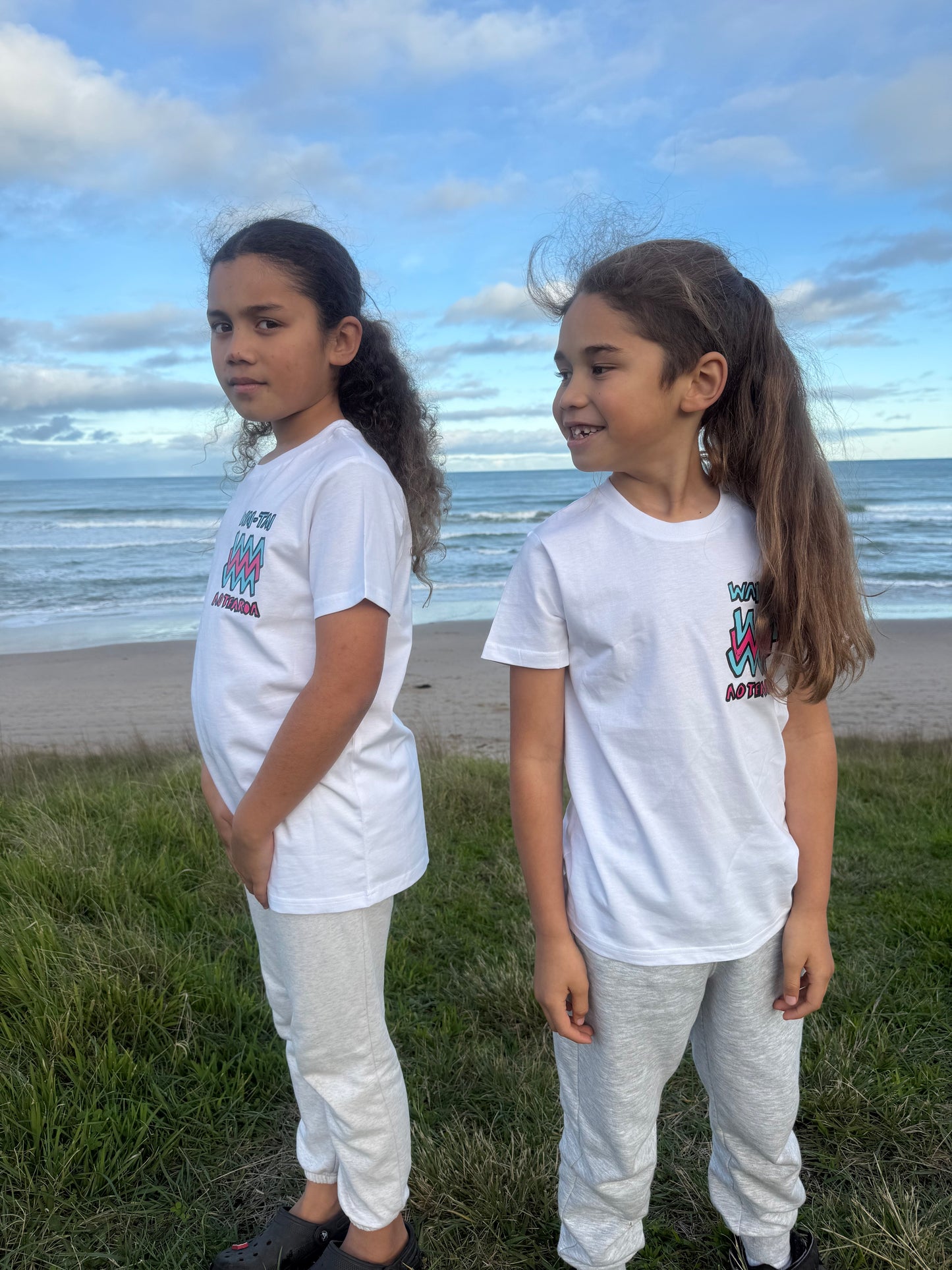 Wai-Tai Kids Mura Maori t-shirt in white from front view on two tamariki at Okltu beach in Gisborne