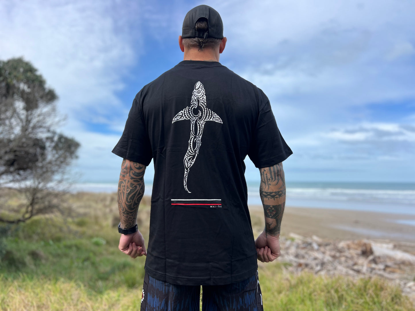Waitai Aotearoa Kaitiaki Tee with a shark on the back in Maori ta moko design on Maia Gibbs at the beach in Gisborne