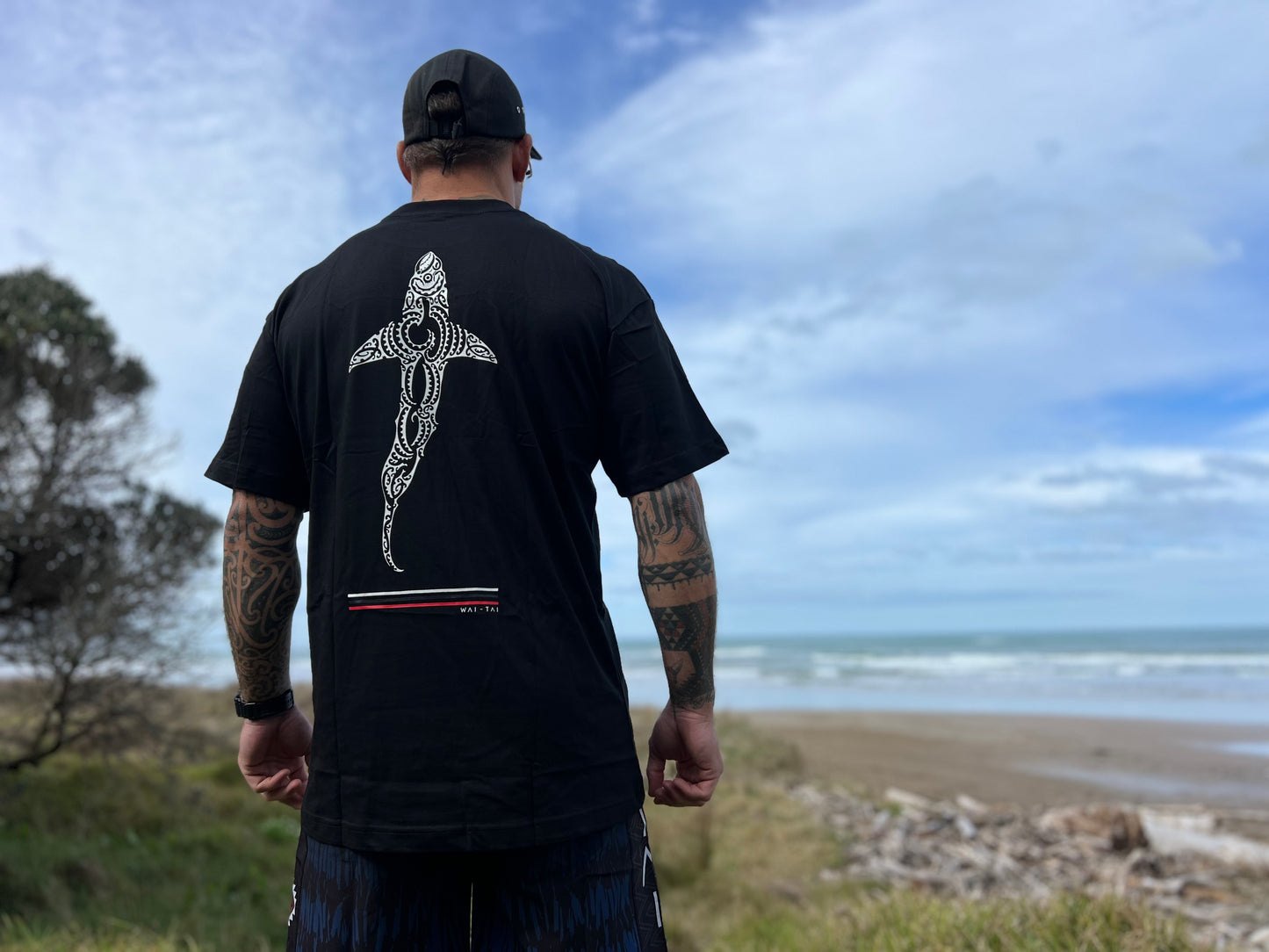 Waitai Aotearoa Kaitiaki Tee with a shark on the back in Maori ta moko design on Maia Gibbs at the beach in Gisborne
