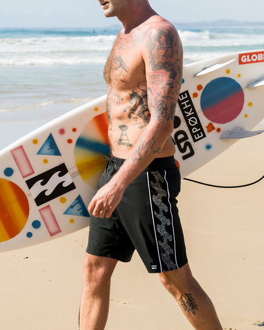 Billabong D Bah Pro 18" Tech Boardshorts being worn by a guy on the beach carrying his surfboard