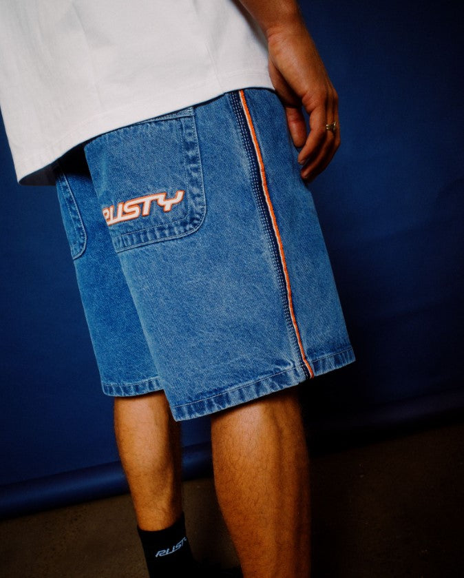 Rusty Flip Daddy 2.0 Denim Shorts in middy blue in lifestyle shot with model wearing white t-shirt on blue background from rear