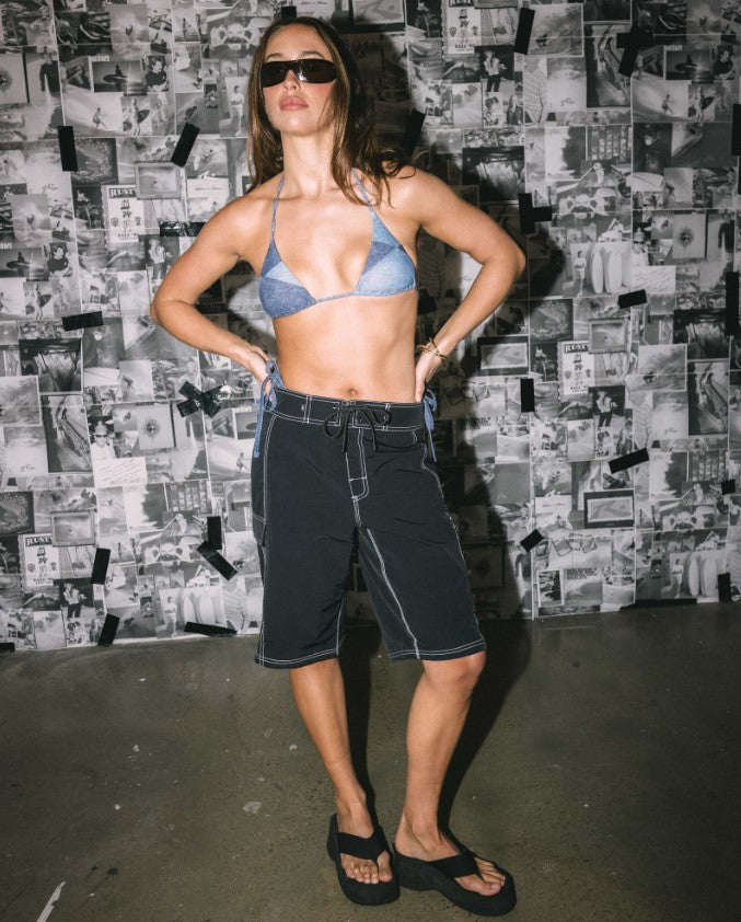Rusty Pick Pocket Womens Boardshorts in black on a model wearing a blue bikini in front of a newspaper collage wall