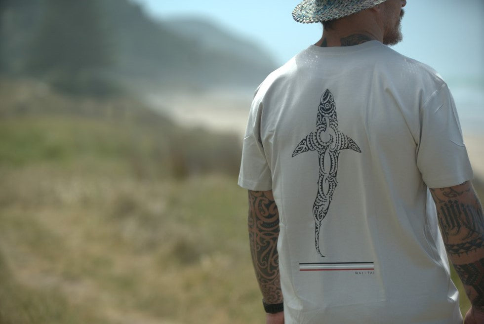 Wai-Tai Mens Kaitiaki Tee in bone colour on Maia Gibbs at Okitu beach