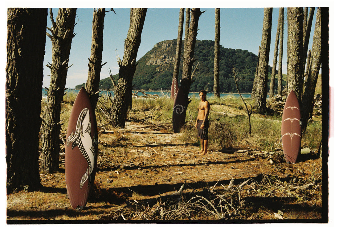 Kehu Butler standing in a forest in his Quiksilver Maori boardshorts with his surfboards standing up around him