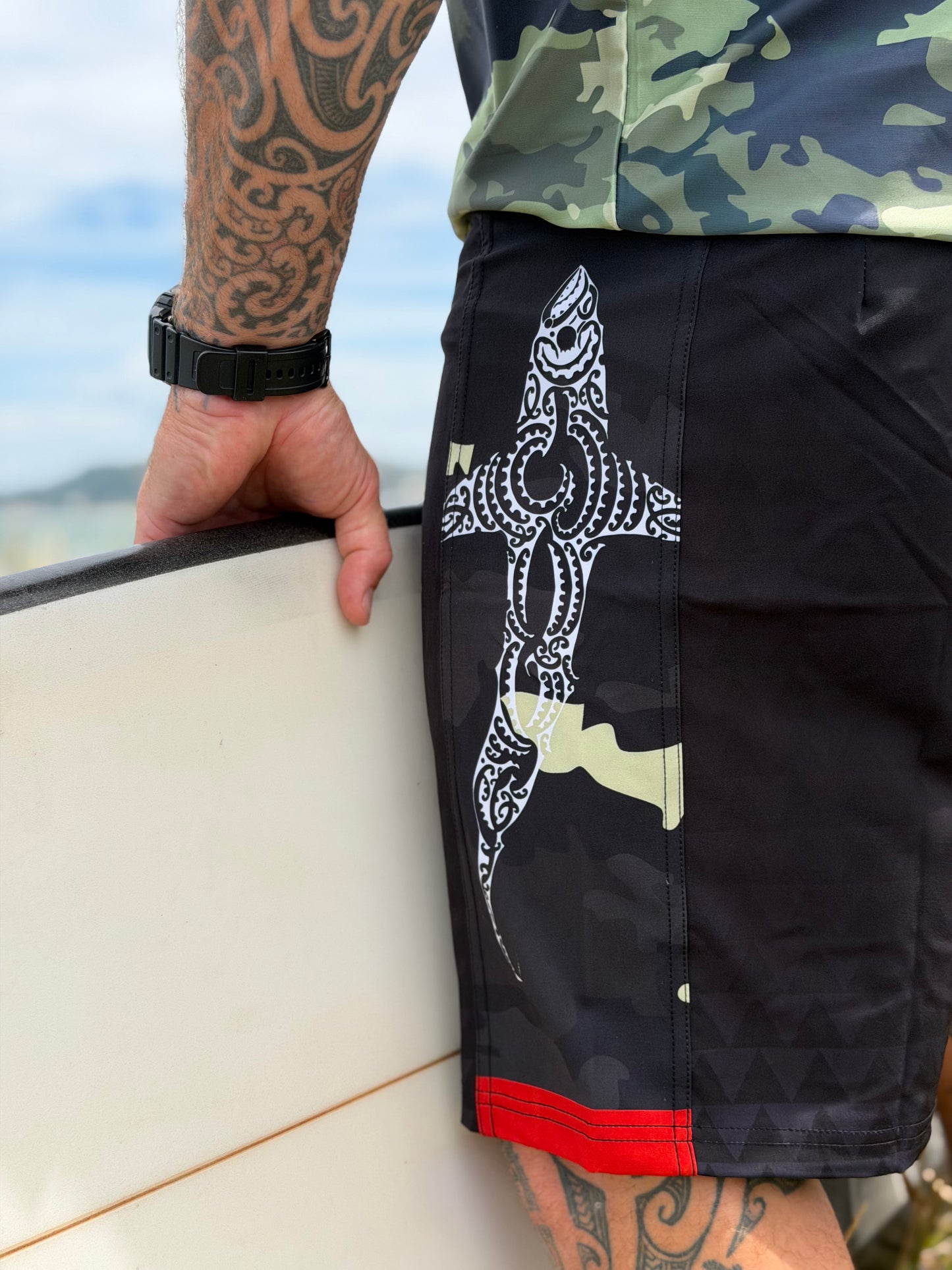 Wai-Tai Aotearoa Kaitiaki Black Camo 19" Tech Maori Design Boardshorts opn Maia Gibbs in camo rashie at Wainui Beach from left side showing shark design