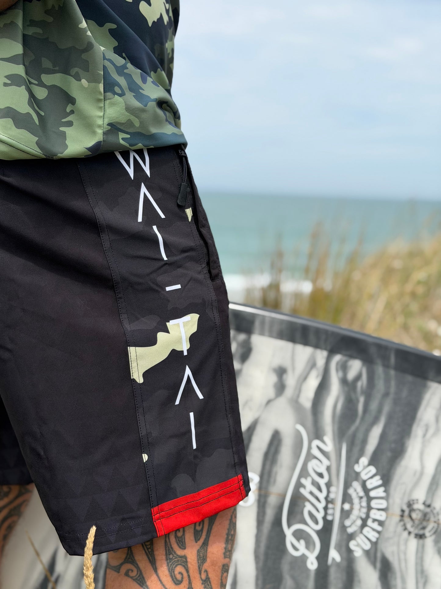 Wai-Tai Aotearoa Kaitiaki Black Camo 19" Tech Maori Design Boardshorts opn Maia Gibbs in camo rashie at Wainui Beach from side