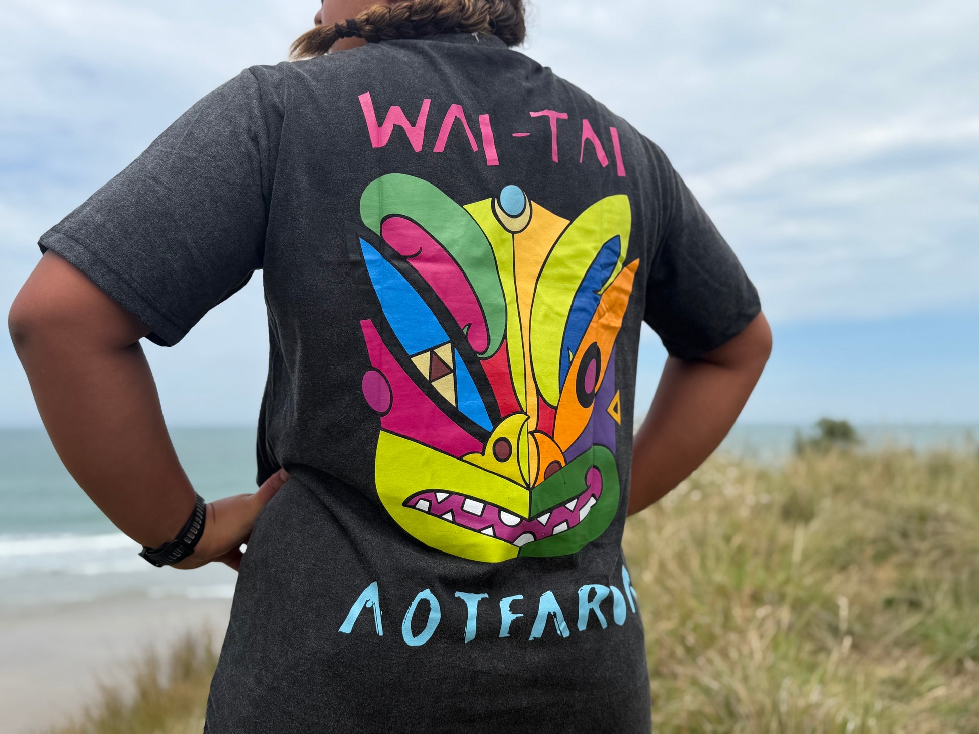 Wai-Tai Aotearoa Ono Whitu Unisex Stonewash Tee from back showing bright coloured tiki design at Okitu beach on Hine Brooking