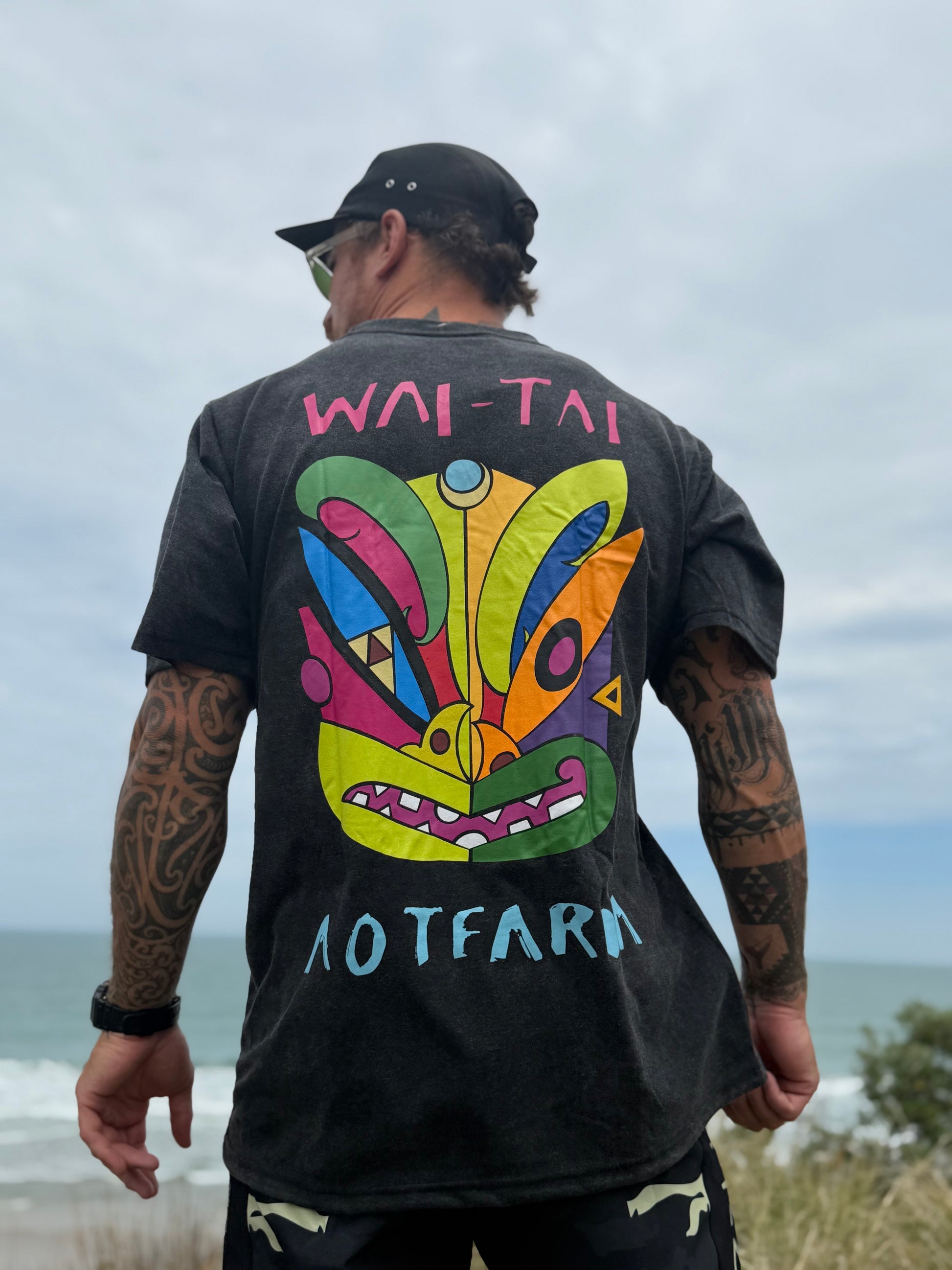Wai-Tai Aotearoa Ono Whitu Unisex Stonewash Tee from back showing bright coloured tiki design at Okitu beach on Maia Gibbs