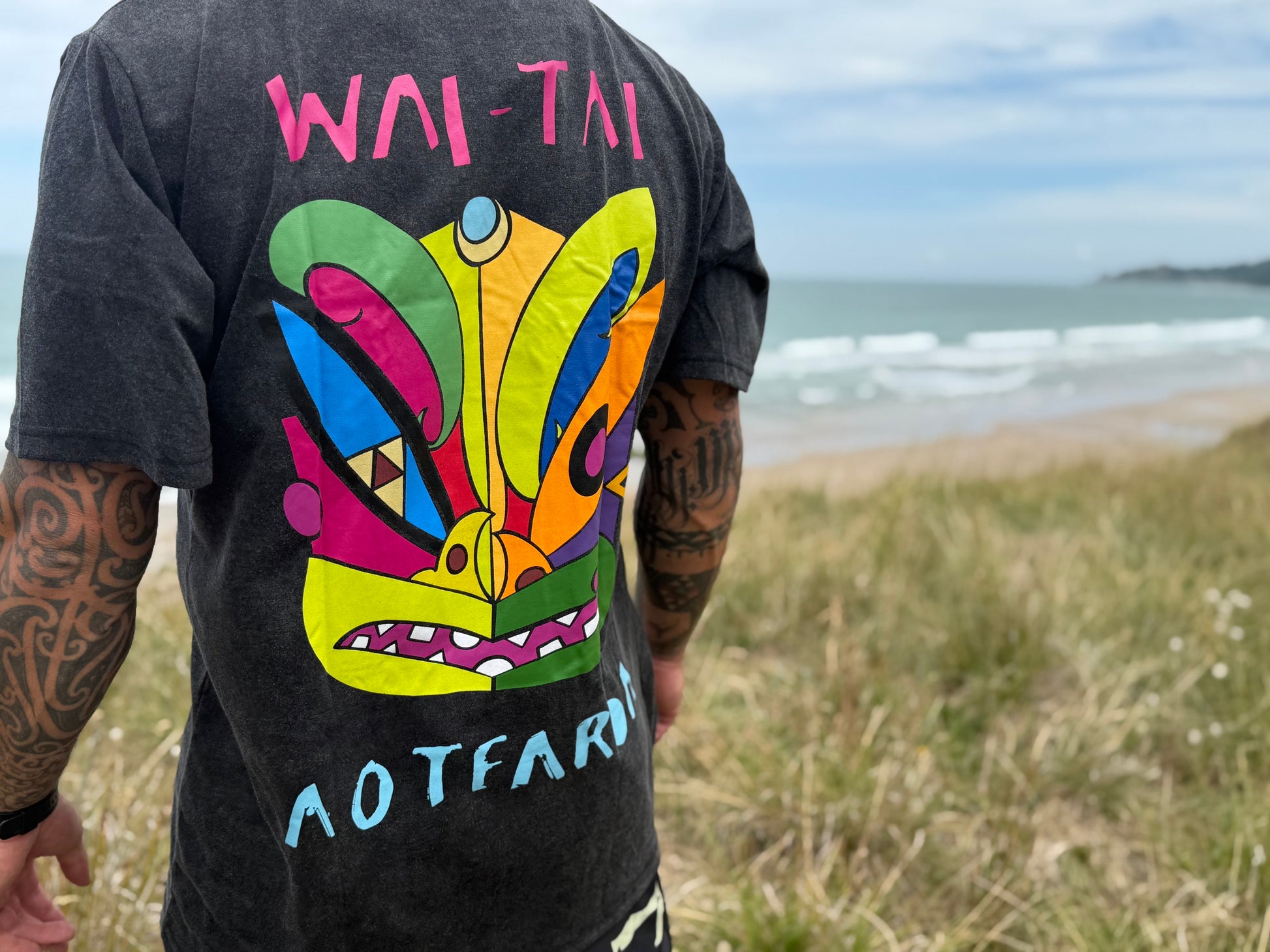 Wai-Tai Aotearoa Ono Whitu Unisex Stonewash Tee from back showing bright coloured tiki design at Okitu beach