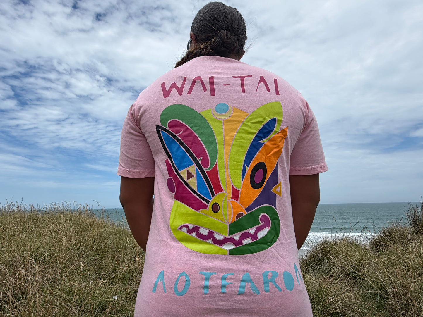 Wai-Tai Aotearoa Ono Whitu Pink Unisex Tee from rear showing bright tiki logo on Hine Brooking at the beach