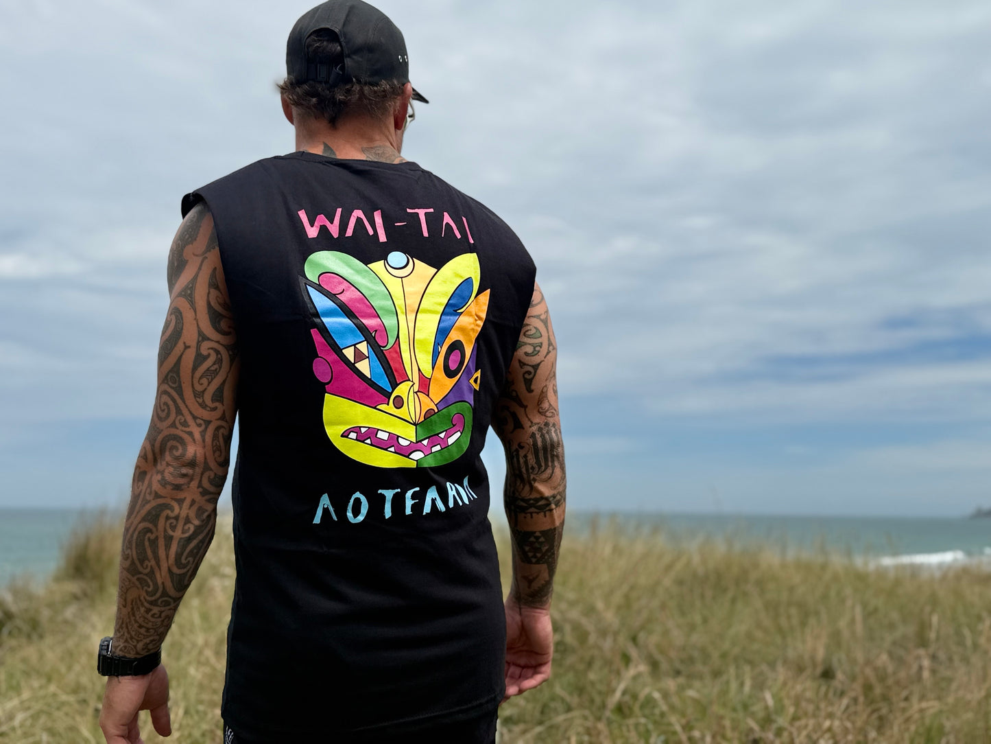 Wai-Tai Aotearoa Ono Whitu Heavyweight Unisex Maori Design Muscle Tee in black on Maia Gibbs at Wainui Beach