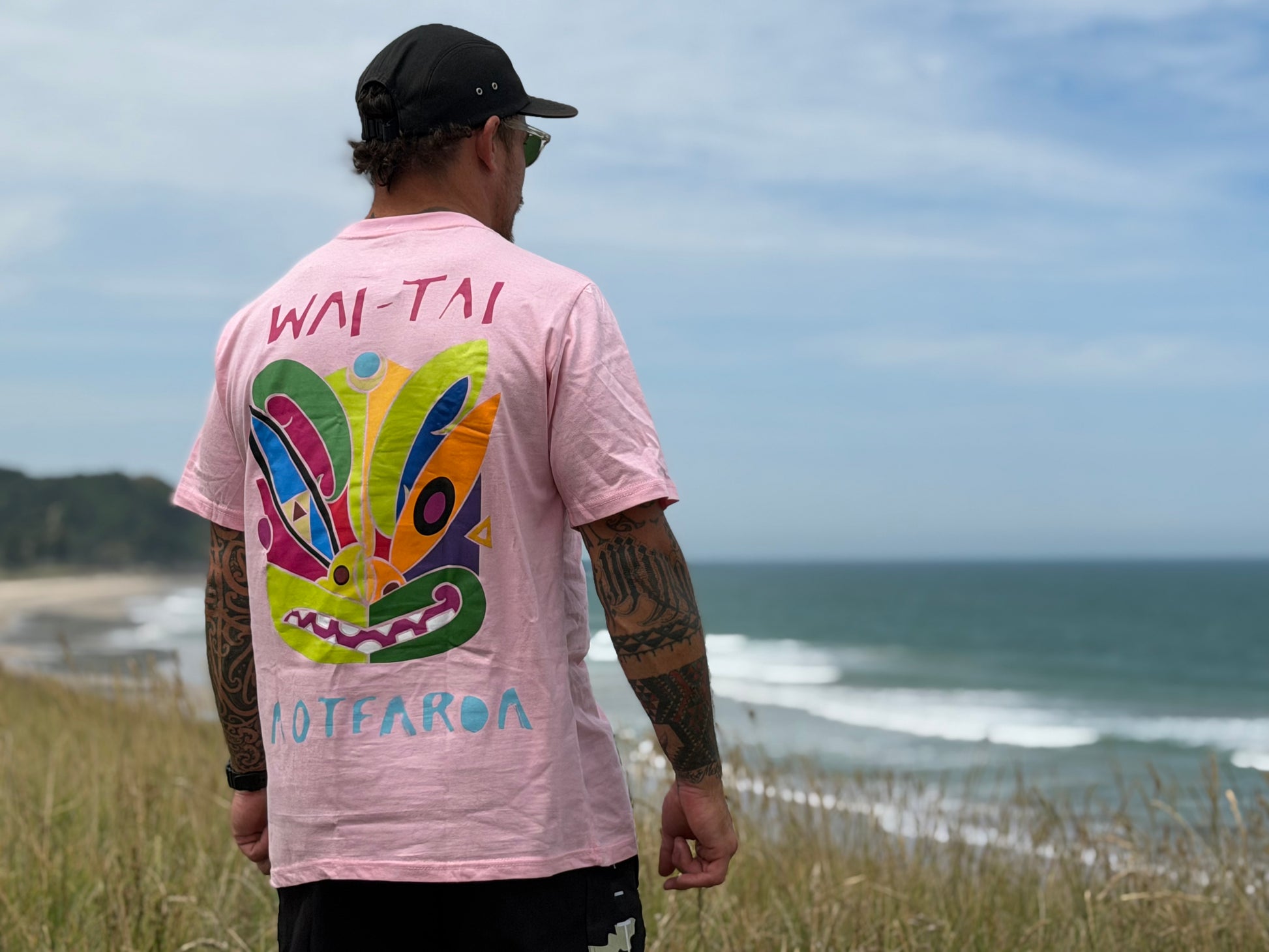 Wai-Tai Aotearoa Ono Whitu Pink Unisex Tee from rear showing bright tiki logo on Maia Gibbs at the beach