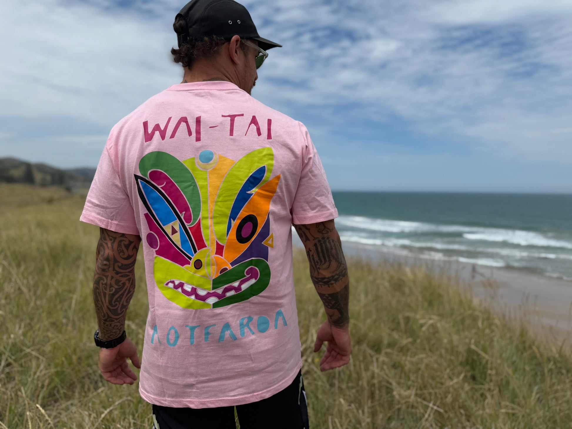 Wai-Tai Aotearoa Ono Whitu Pink Unisex Tee from rear showing bright tiki logo on Maia Gibbs at the beach