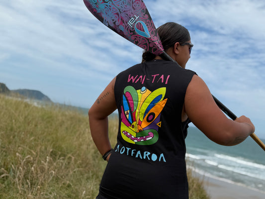 Wai-Tai Aotearoa Ono Whitu Heavyweight Unisex Maori Design Muscle Tee in black on Hine Brooking carrying a waka paddle at Wainui Beach