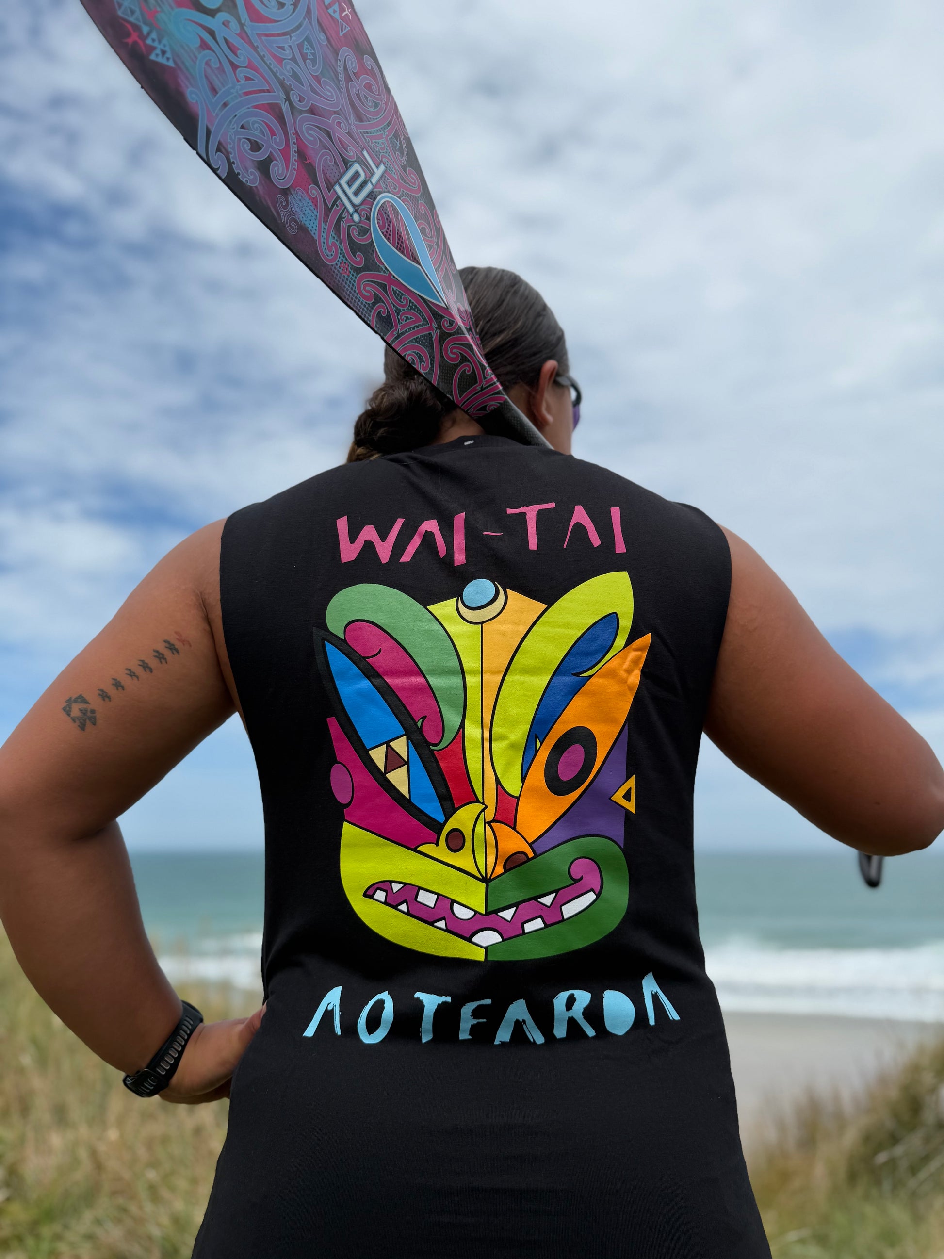 Wai-Tai Aotearoa Ono Whitu Heavyweight Unisex Maori Design Muscle Tee in black on Hine Brooking carrying a waka paddle at Wainui Beach