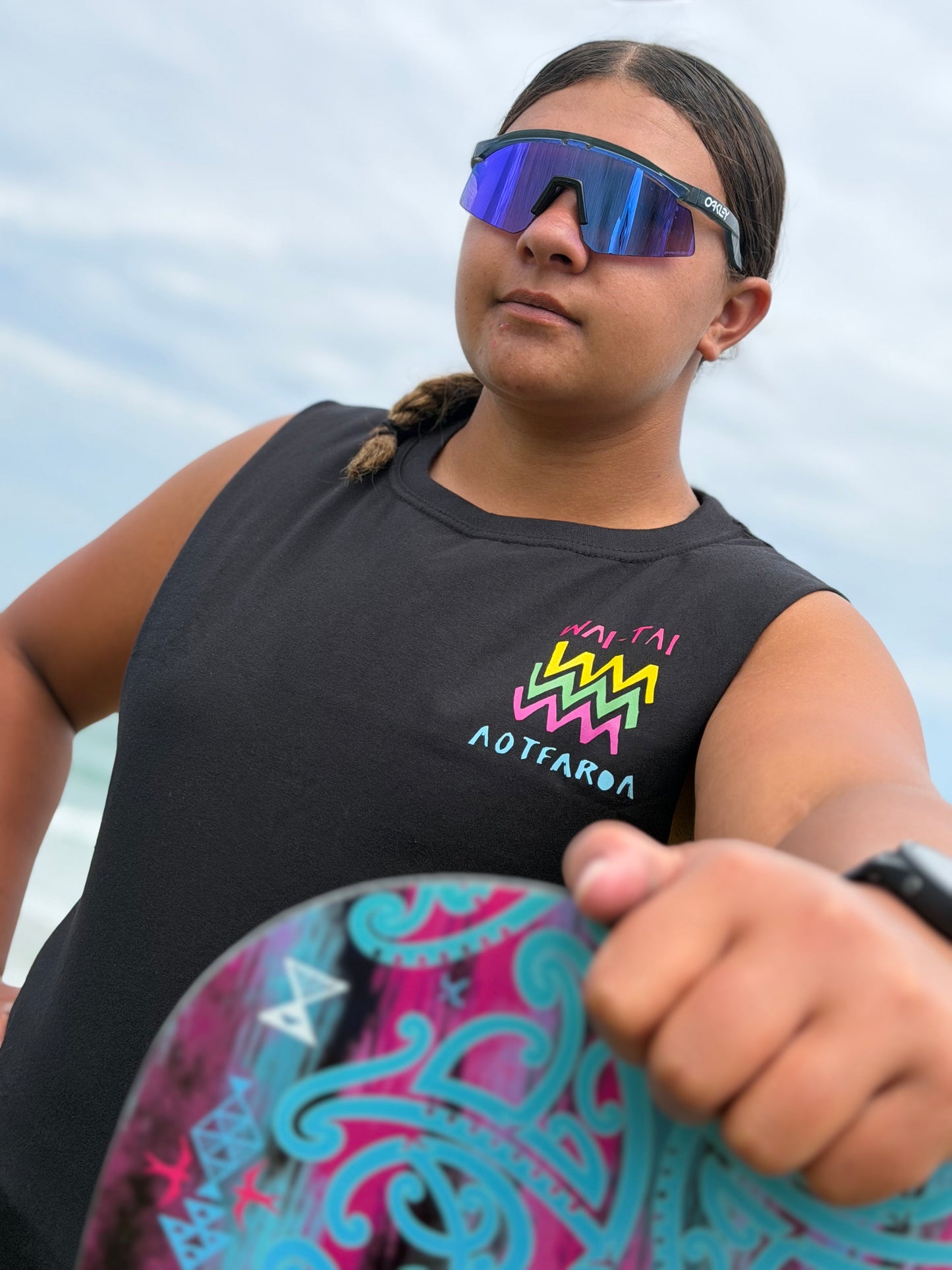 Wai-Tai Aotearoa Ono Whitu Heavyweight Unisex Maori Design Muscle Tee in black on Hine Brooking carrying a waka paddle at Wainui Beach