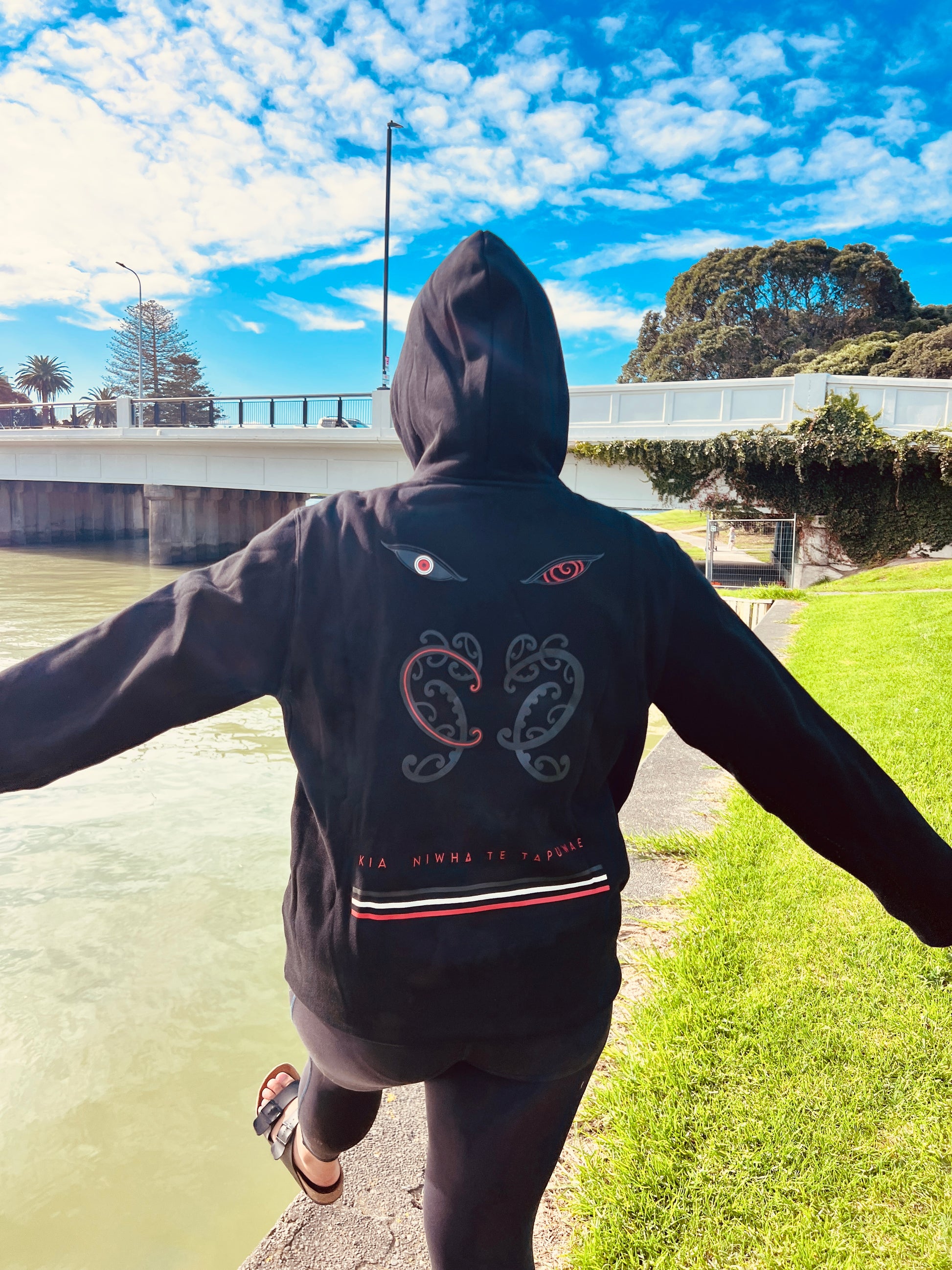 Wai-Tai Tipua Blackout Heavyweight Maori Design Unisex Hoodie in black from the rear by river in Gisborne