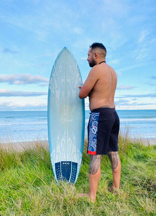 Wai-Tai Aotearoa Taniwha 4 Way Stretch Maori Boardshorts on Daniel Procter holding a Maori design surfboard from rear at Okitu beach