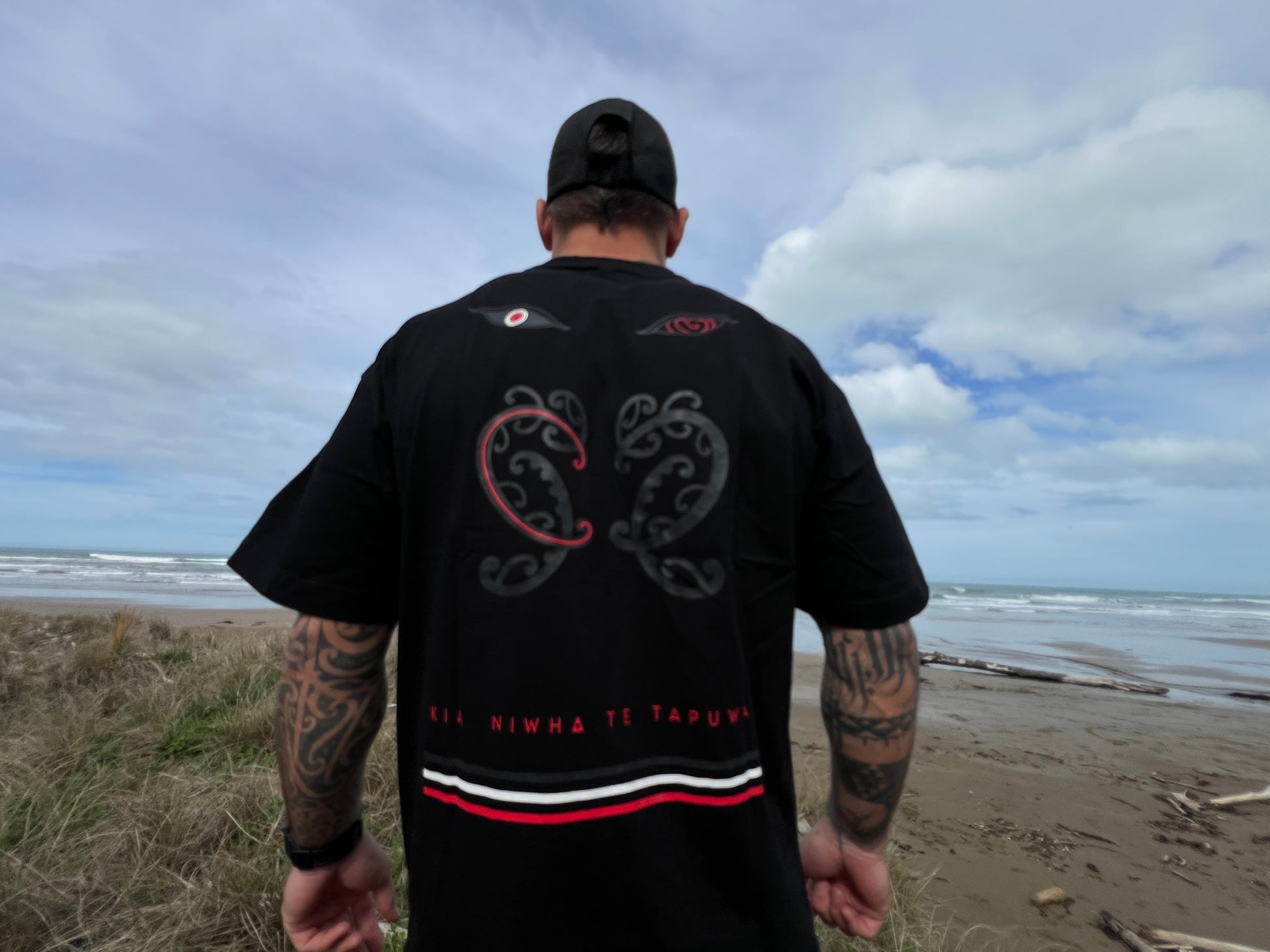 Wai-tai Tipua Heavyweight Blackout Tee showing Tanagaroa's face in a blackout style on Maia Gibbs from rear view at the beach