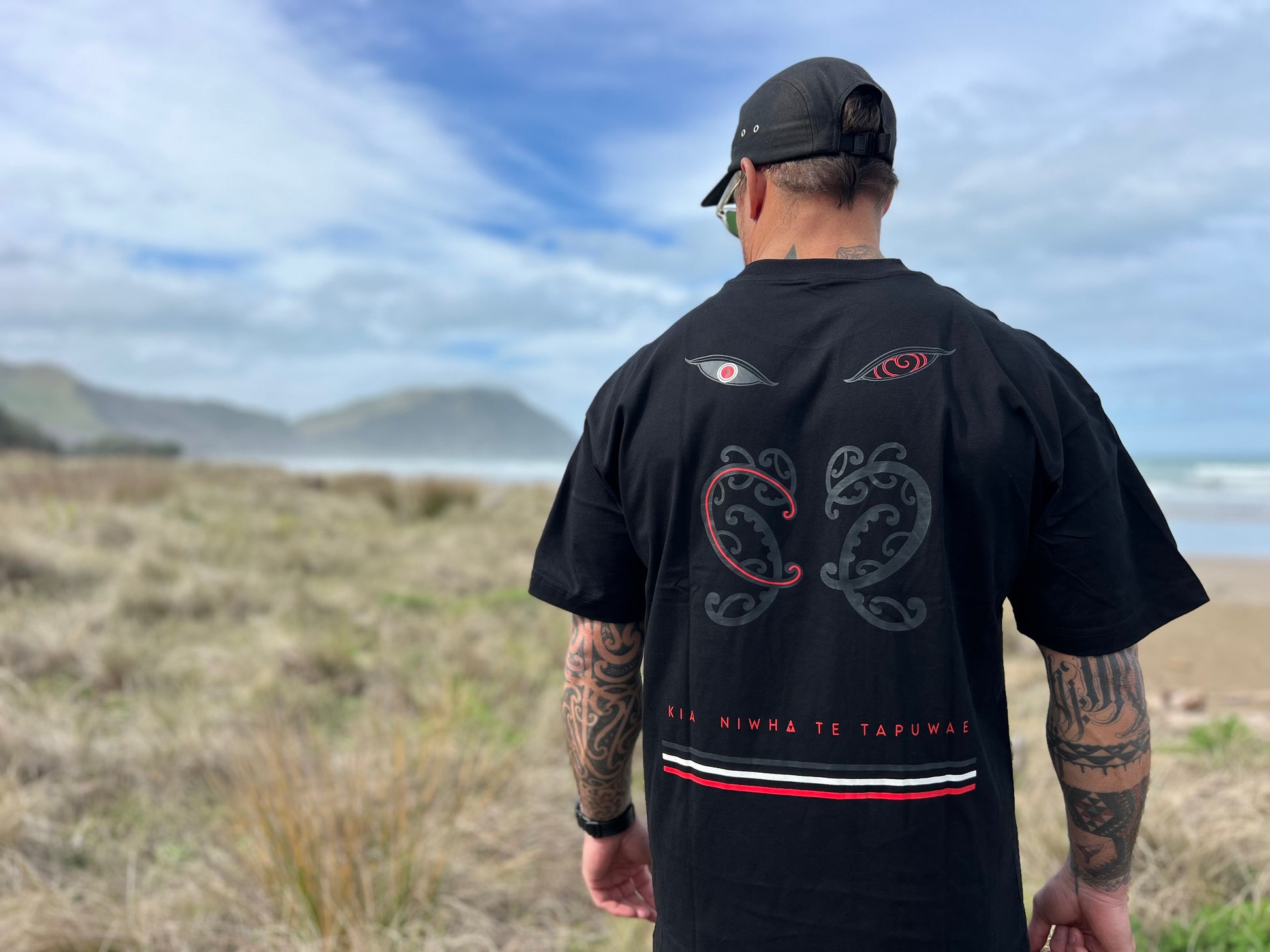 Wai-tai Tipua Heavyweight Blackout Tee showing Tanagaroa's face in a blackout style on Maia Gibbs from rear view at the beach