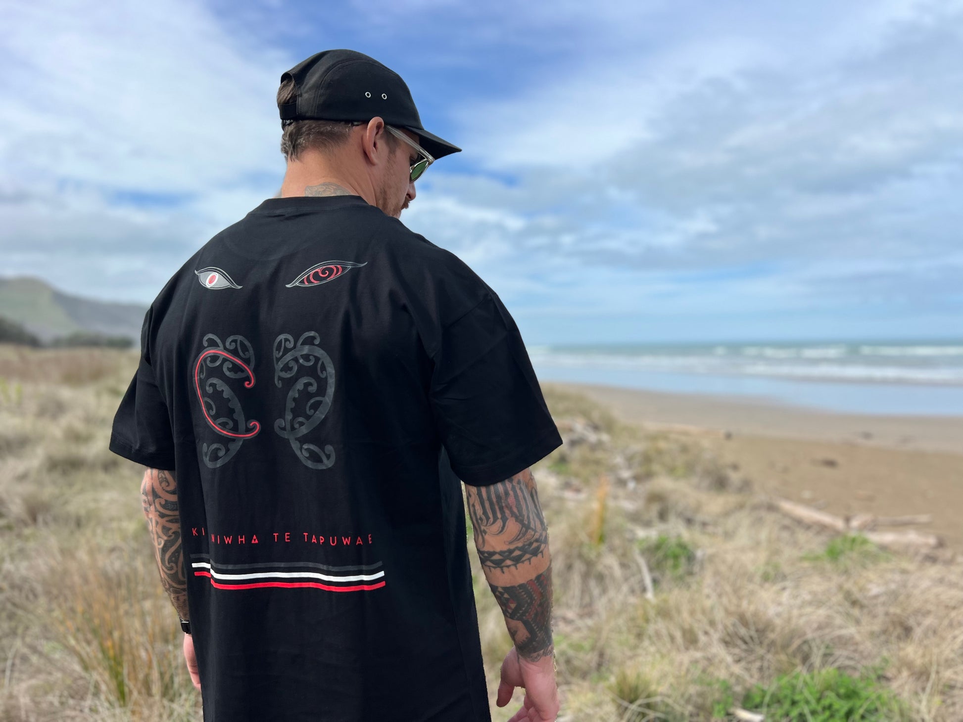 Wai-tai Tipua Heavyweight Blackout Tee showing Tanagaroa's face in a blackout style on Maia Gibbs from rear view at the beach