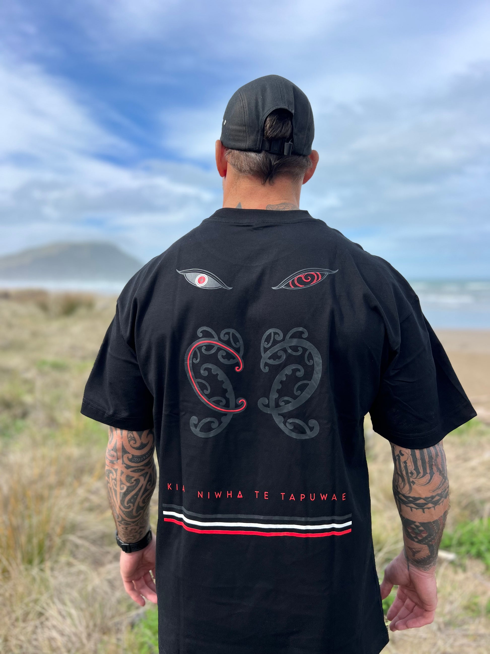 Wai-tai Tipua Heavyweight Blackout Tee showing Tanagaroa's face in a blackout style on Maia Gibbs from rear view at the beach