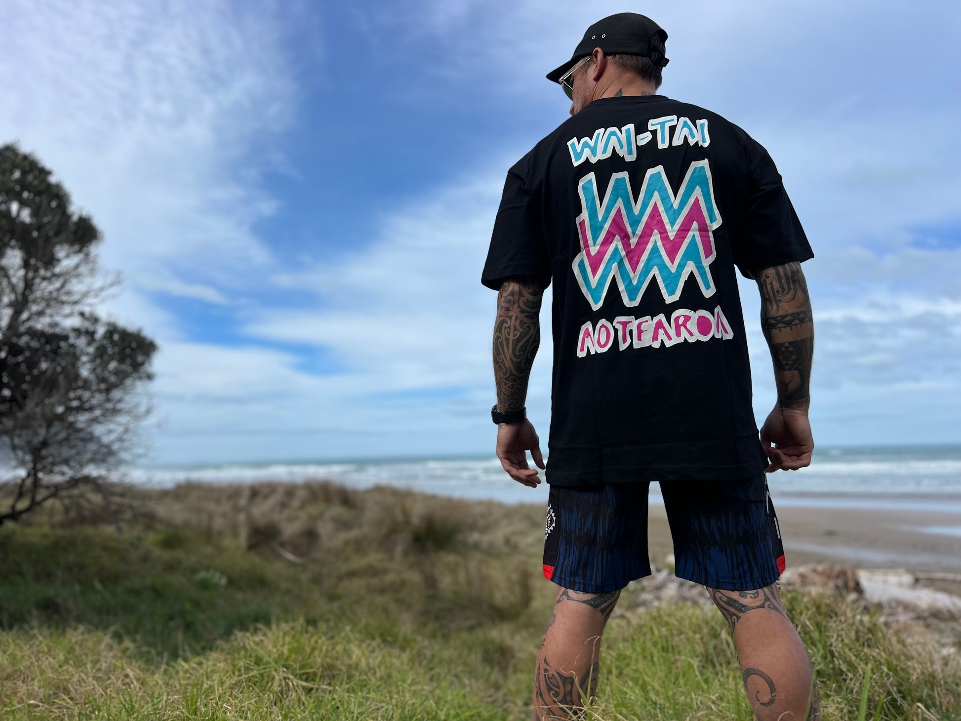 Wai-Tai Aotearoa Mura heavyweight Maori t-shirt in black with flouro print on Maia Gibbs from the back view wearing Wai-Tai piu boardshorts