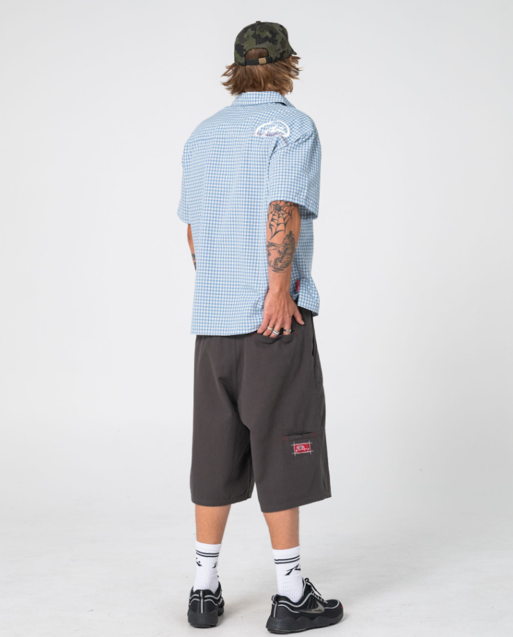 Rusty Stasher 25" Baggy Canvas Walkshorts in carbon colourway on model in blue checked shirt and Rusty cap from the back