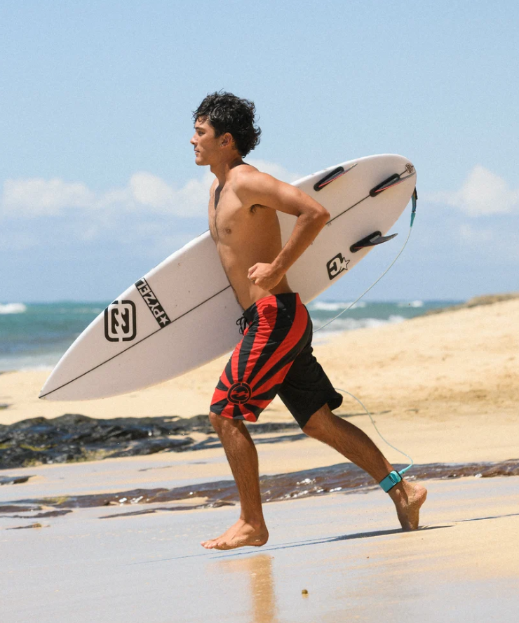 Billabong A.I Rising Sun Pro Boardshorts in black and red on surfer running down the beach with his Pyzel surfboard