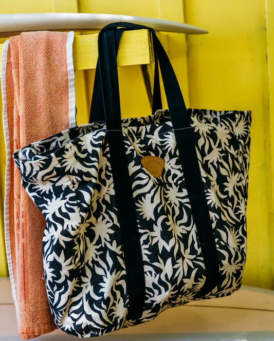 Billabong Poppins Playa Tote Bag in black and white floral print hanging from a surfboard with yellow background