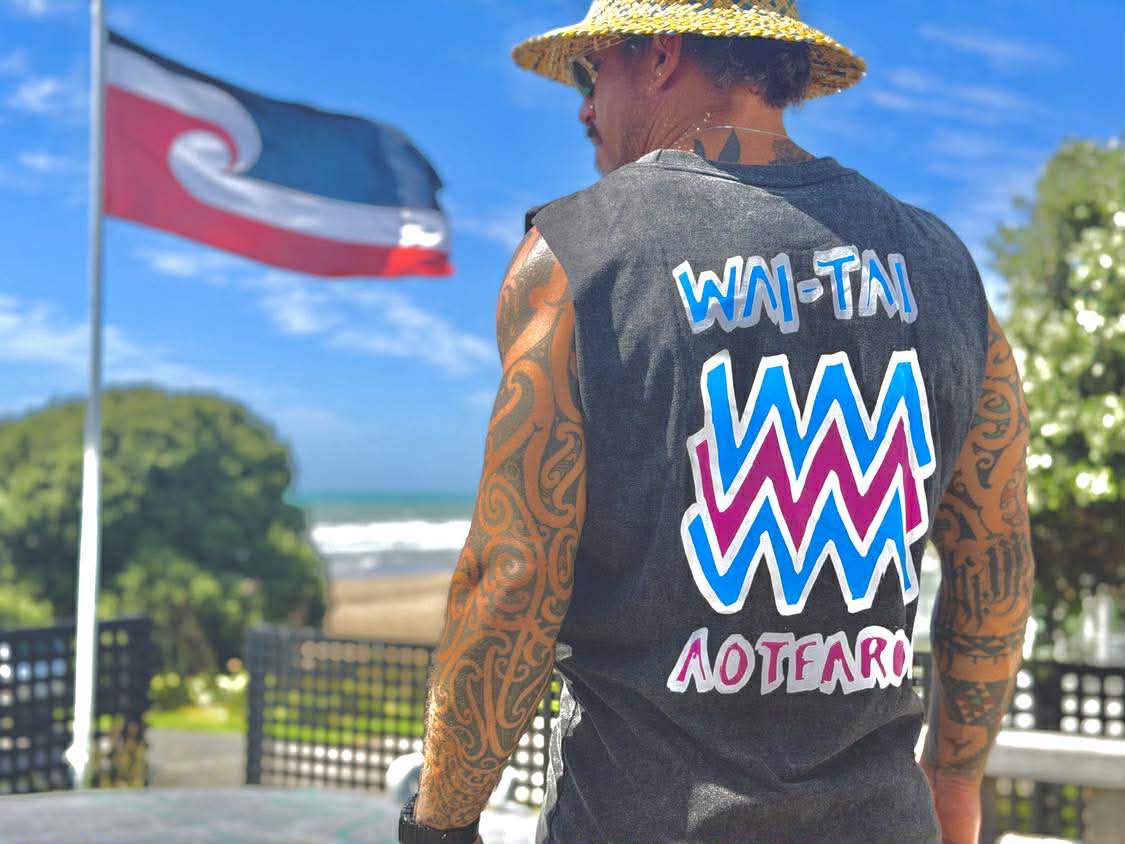 Wai-Tai Mura Stonewash Heavyweight Unisex Muscle in black stonewash with flouro logo on Maia Gibbs with Tino Rangatiratanga flag in background