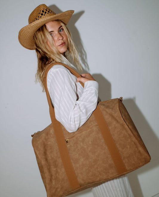 Blonde woman in white dress holding the Rusty Aria Suede Weekender Bag in tan colourway