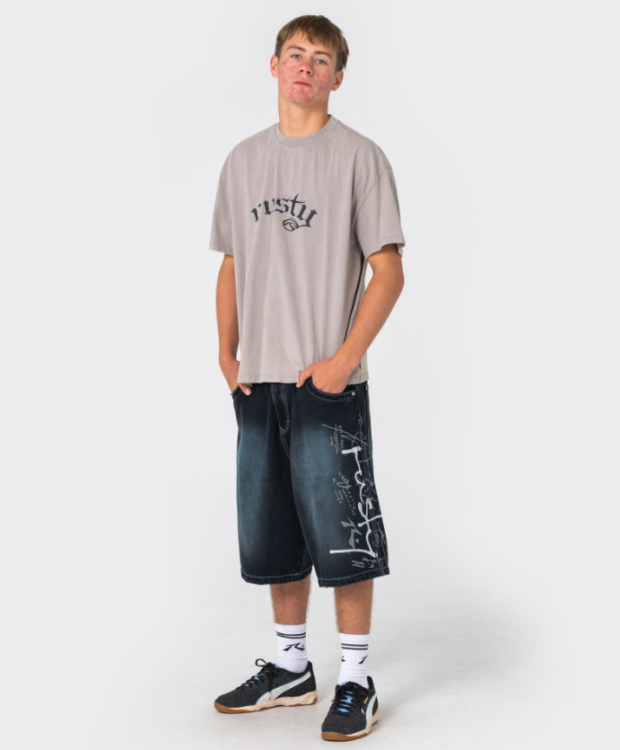 Rusty Kurban Spray Baggy 25" Denim Shorts in blackout petrol colourway on model in beige coloured Rusty tee from front slight left side to show large Rusty side print