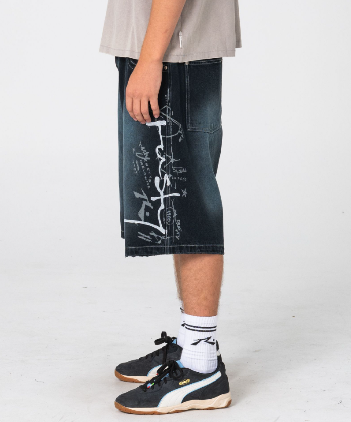 Rusty Kurban Spray Baggy 25" Denim Shorts in blackout petrol colourway on model in beige coloured Rusty tee from side showing large Rusty print