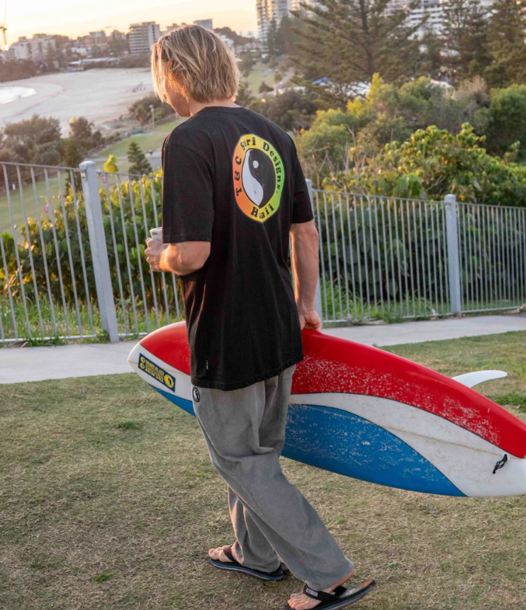 T & C OG Logo Men's T-Shirt in black with rasta print on man holding red, white and blue surfboard