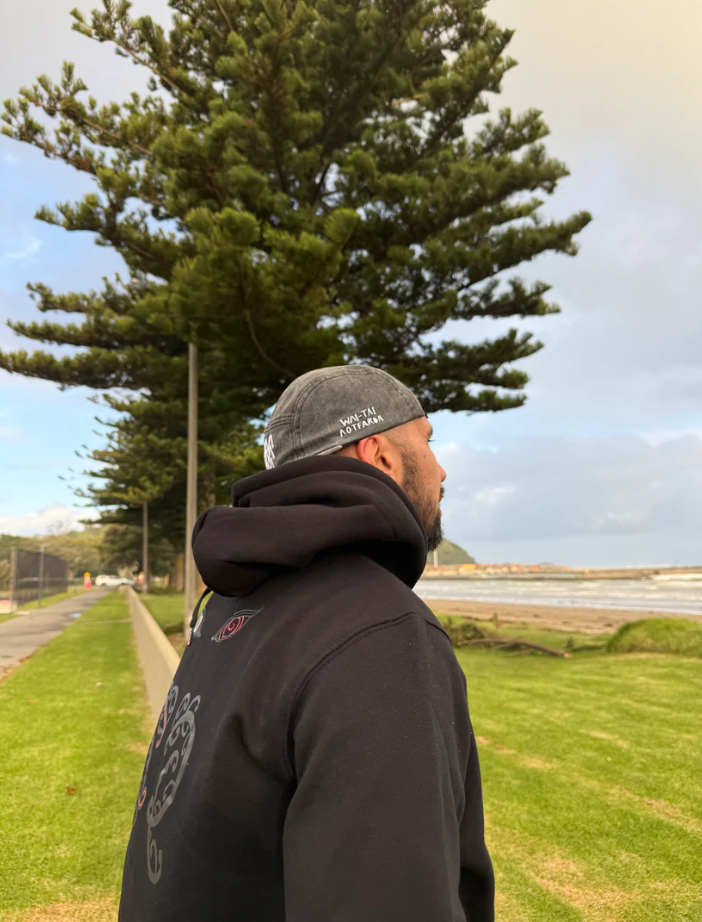 Wai-Tai Aotearoa Tohu Denim 5 Panel Unisex SB Maori Design Cap in black acid colourway on Daniel Procter at the beach in Gisborne