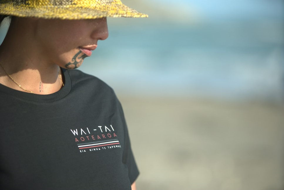 Wai-Tai Womens Tohu Tee in black showing front logo at Kaiti Beach in Gisborne